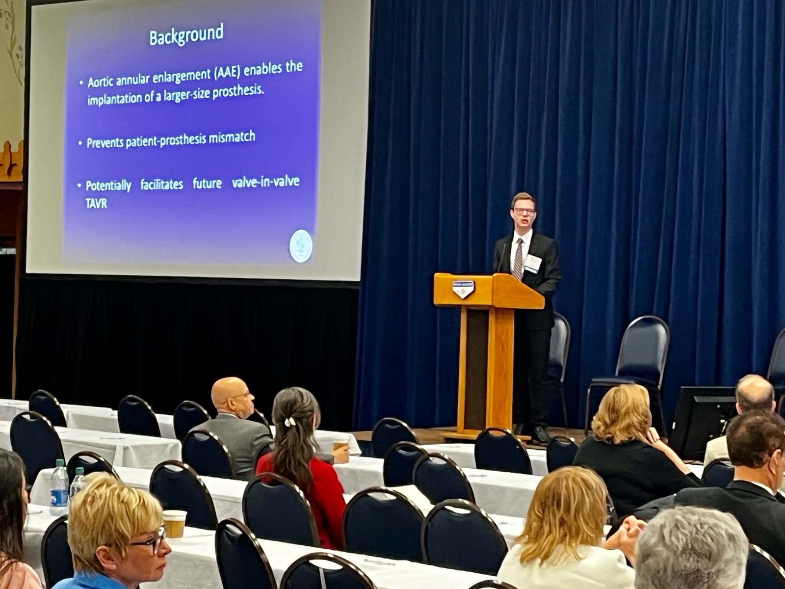 Dimitrios Magouliotis lecturing from a podium to an audience, beside a slide on aortic annular enlargement.